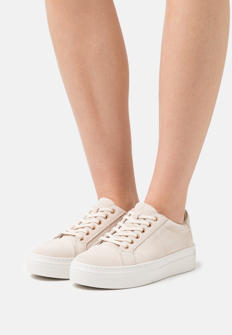 Světle béžové semišové tenisky s kulatou špičkou, bílou platformovou podrážkou, zlatými kovovými očky a plochými tkaničkami.