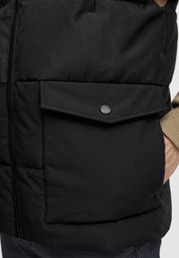 Gilet imbottito nero con chiusura a zip, dotato di una grande tasca frontale chiusa da un bottone a pressione nero. Tessuto testurizzato e design strutturato.