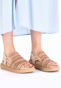 Bunte gewebte Sandalen mit einem atmungsaktiven Textil-Obermaterial, einem beigen Zehenriemen und Knöchelunterstützung, auf einer hellbraunen Gummisohle.