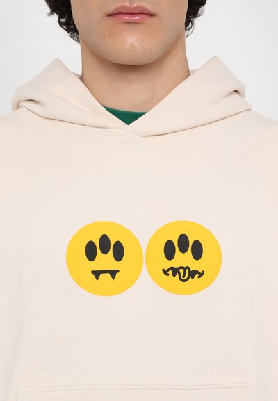 Light beige hoodie featuring two large yellow circles with black playful character designs in the center. Soft fabric with a relaxed fit.