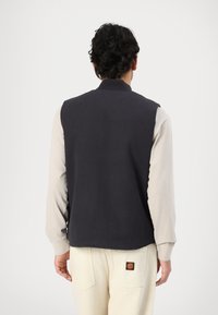 Sleeveless dark navy vest made from textured fabric, featuring a ribbed collar. Worn over a long-sleeve light-colored shirt and beige pants.