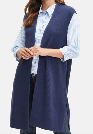 Kobieta ubrana w bezrękawnik z granatowego dzianiny na jasnoniebieskiej i białej koszuli w paski z guzikami oraz ciemne dżinsy, z rękami złożonymi na wysokości talii.