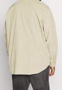 Beżowa koszula z długim rękawem o luźnym kroju, z mankietami na guziki, zaokrąglonym dołem i miękką fakturą. Noszona z ciemnoszarymi jeansami.