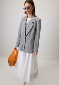 Pilkos smailių raštų švarkas su dideliu apykaklė, derinamas su balta maksi suknele. Laikoma oranžinė tote kuprinė, o ant kojų lengvi sportbačiai ir saulės akinius.