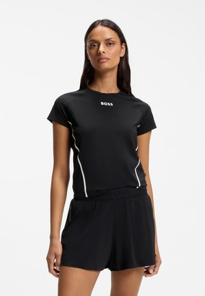 Frau trägt ein schwarzes Kurzarmtop mit weißem "BOSS"-Logo und passende schwarze Shorts, steht vor einem schlichten weißen Hintergrund.