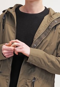 Olive zelená bunda se zapínáním na zip vpředu, nastavitelnou kapucí a stahovacím páskem v pase. Pod ní černý strukturovaný svetr, ruce sepnuté před tělem.