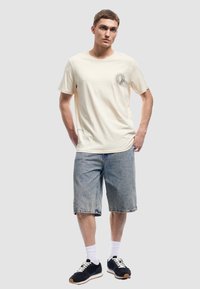 Joven con una camiseta de crema con estampado de sol, pantalones cortos de mezclilla hasta la rodilla, calcetines blancos y zapatillas negras, de pie con las manos en los bolsillos.