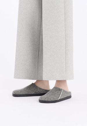 Piedi con pantofole slip-on grigie a motivo spina di pesce con suole nere, indossate con pantaloni grigio chiaro a gamba larga, parzialmente visibili, su sfondo bianco.