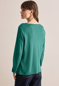 Teel pitkähihaine paita, joka on valmistettu pehmeästä materiaalista, leveällä kaula-aukolla ja rentoa istuvuutta. Takana on kaareva helma ja yksinkertainen muotoilu.