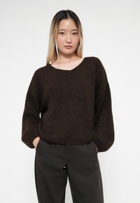 Hnědý pletený svetr s výstřihem do V a volnými dlouhými rukávy. Texturovaná látka a uvolněný střih. Kombinován s tmavými rovnými kalhotami.