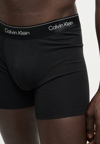 Άνδρας φοράει μαύρα μπόξερ Calvin Klein, δείχνοντας τη μέση και το επάνω μέρος των μηρών, με ορατή την επωνυμία στη μέση.