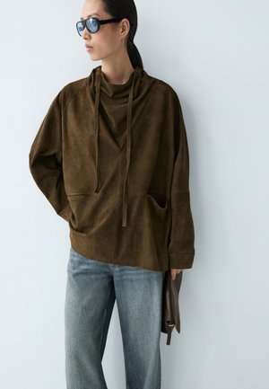 Frau in dunkelbraunem, locker sitzendem Hoodie und hellblauen Jeans, die schwarze Sonnenbrillen trägt und eine dunkelbraune Clutch vor einer schlichten hellen Wand hält.
