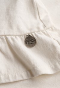 Crème-kleurige stof met een gerimpelde zoom, voorzien van een ronde, zilveren label met de opdruk "SuperDry Brand" in decoratieve tekst.