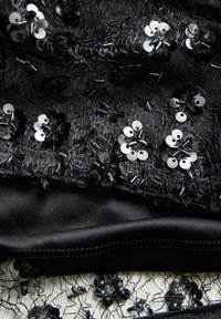 Melna mežģīņu auduma ar sequins un pērlītēm ziedu rakstos, slāņotā ar gludu melnu materiālu apakšā. Tekstūrēts un detaļām bagāts dizains.