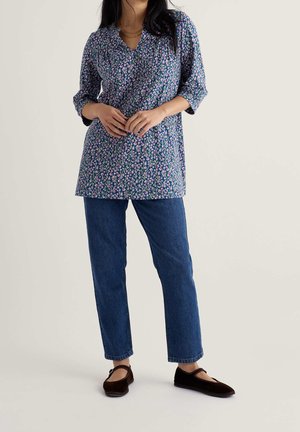 Frau mit einer blauen, geblümten Bluse mit langen Ärmeln, blauen Jeans und dunkelbraunen Mary Jane Schuhen, stehend vor einem schlichten Hintergrund.