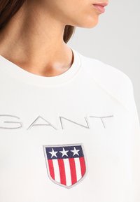 Mujer vistiendo una camisa blanca con "GANT" bordado y un emblema de escudo que presenta tres estrellas blancas sobre franjas rojas y blancas.
