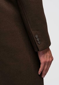 Miehen käsi tummanruskean villatakin kalvon vieressä, jossa on kolme mustaa nappia hihansuussa, vaaleaa taustaa vasten.