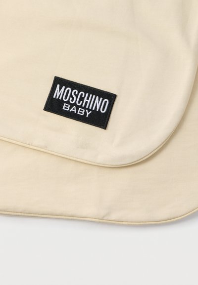 Μπεζ βαμβακερή παιδική κουβέρτα με στρογγυλεμένες γωνίες, με μαύρη κεντημένη ετικέτα με το κείμενο "MOSCHINO BABY" σε λευκό. Μαλακή υφή.