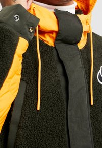 Fleecfodrad jacka med en orange skalfärg, svarta detaljer och en strukturerad yta, med dragsko och knappdetaljer.