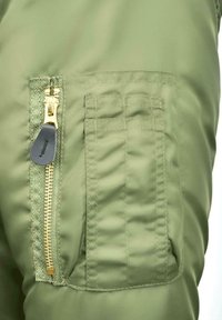 Alyvuogių žalios spalvos striukės audinys su šonine kišene, turinčia auksinį užtrauktuką ir juodą traukimo juostelę. Lygi tekstūra su išsamiais siūlėmis.