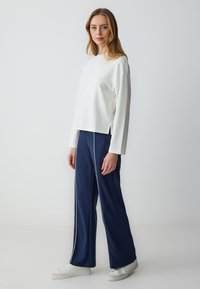 White long-sleeve top with subtle embellishments and side slits, paired with navy wide-leg pants featuring white piping, and white sneakers.