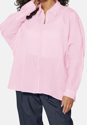 Mujer que lleva una camisa rosa pálido de manga larga y suelta abotonada y pantalones gris oscuro, con una mano tocando su cabello.