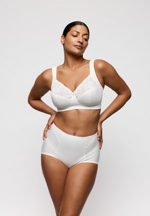 Vrouw poseert in een witte beha met kanten accenten en high-waisted slip tegen een effen lichte achtergrond, handen rustend op haar heupen.