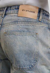 Jasnoniebieskie dżinsy z wyblakłą fakturą, z skórzaną metką "2Y STUDIOS" na tylnym pasku oraz standardowymi kieszeniami.