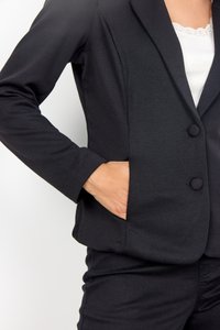Schwarzer Blazer mit zwei Knöpfen, einem Reverskragen und Taschen. Der Stoff wirkt glatt und dehnbar und wird mit einem weißen Hemd mit Spitzenbesatz darunter kombiniert.