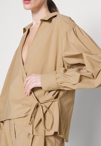 Beige wrap-paita pitkäpuffeilla hihoilla, syvällä V-kaula-aukolla ja sivusiteellä. Valmistettu sileästä kankaasta, jossa on rento istuvuus.