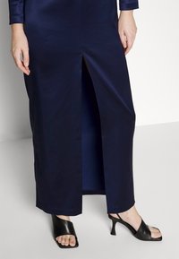 Navy satin dress with a thigh-high slit, featuring long sleeves and a smooth texture, paired with black open-toe high-heeled mules.