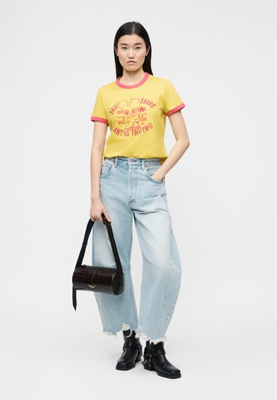 Νεαρή γυναίκα που φοράει κίτρινη μπλούζα με τύπωμα, ανοιχτό μπλε παντελόνι φαρδύ με σκισίματα, μαύρες μπότες και κρατάει μαύρη κυλινδρική τσάντα.