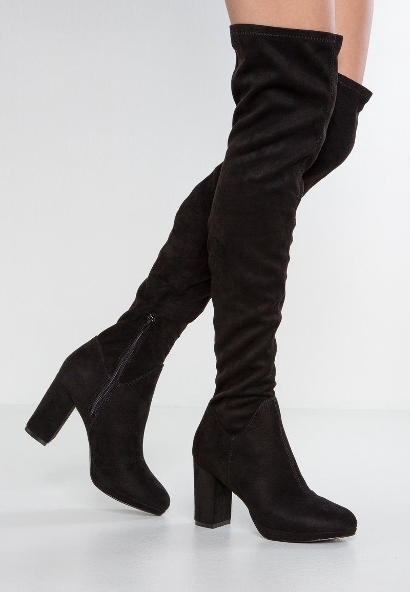 Mustat nubukista valmistetut polven yli ulottuvat saappaat, joissa on paksut korot ja sivuvetoketjut, jalassa ristittyjen jalkojen päällä vaaleaa taustaa vasten.