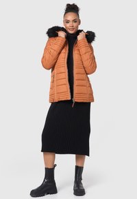 Puffer jacket in burnt orange with a detachable black fur hood, zip closure, and quilted texture, paired with a black ribbed midi dress and black boots.