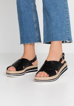 Pieds portant des sandales compensées noires tissées avec des brides à la cheville et un jean bleu à ourlet effiloché sur un sol blanc.