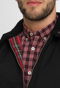 Veste zippée noire avec un col texturé et une chemise à carreaux rouges en dessous. L'intérieur présente un motif rayé coloré. Quincaillerie en argent visible.