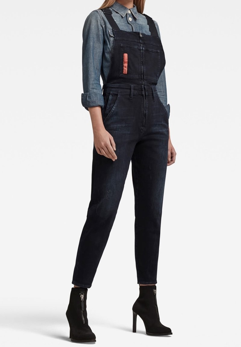 Femme portant une salopette en jean noire avec une fermeture éclair à l'avant, une chemise grise à boutons aux manches retroussées et des bottines à talons noires avec des fermetures éclair.
