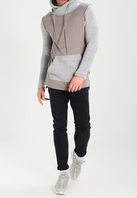 Felpa con cappuccio bicolore in grigio chiaro e beige, dotata di tasca a marsupio e maniche texturizzate. Abbinata a pantaloni neri e sneakers chiare.