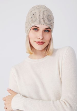 Beige gebreide muts met verticale ribbels, gestructureerde textuur en een stevige pasvorm. Draagt bij een lichtgekleurde trui met een ronde halslijn.