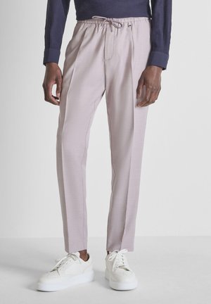 Uomo che indossa pantaloni rosa chiaro con coulisse e scarpe da ginnastica bianche, in piedi davanti a uno sfondo bianco semplice.