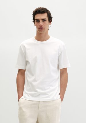 Junger Mann mit dunklem Haar, der ein einfaches weißes kurzärmeliges T-Shirt und hellbeige Hosen trägt, steht mit den Händen in den Taschen.