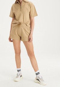 Beige bavlněné polo tričko s krátkým rukávem a polovičním zipem, odpovídající elastické šortky a bílé tenisky s černými akcenty a pruhy na ponožkách.