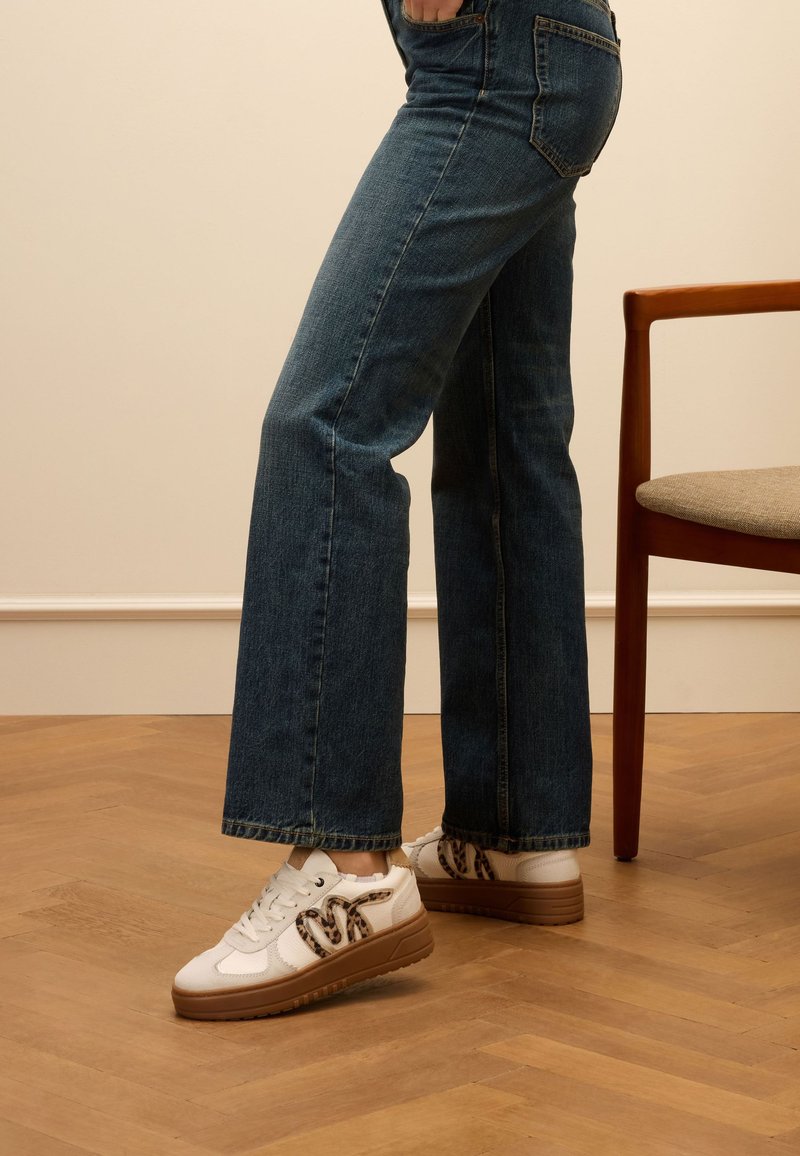 Weiße Sneakers mit brauner Sohle und Akzenten in Leopardenmuster, kombiniert mit weit geschnittenen blauen Jeans. Der Hintergrund zeigt einen Holzfußboden und einen Stuhl.