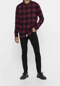 Punainen ja musta ruudullinen pitkähihainen paita, jossa napitus ja terävä kaulus, yhdistetty mustiin kapealahkeisiin housuihin ja mustiin kenkiin.