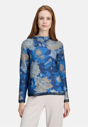 Vrouw in een blauw en beige gebloemd gebreid sweater met lange mouwen en lichte broek, staand tegen een witte achtergrond.