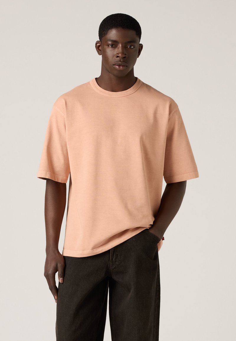 Junger Mann steht mit einer Hand in der Hosentasche, trägt ein pfirsichfarbenes oversized T-Shirt und schwarze Hose vor neutralem Hintergrund.