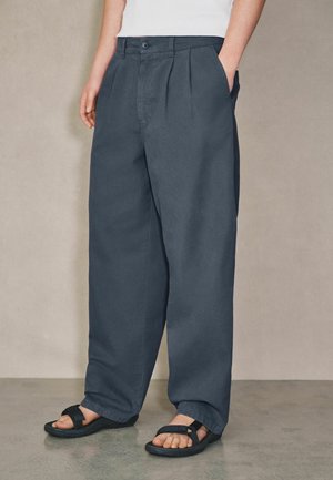 Person wearing loose dark blue pants with front pleats and black sandals, hands in pockets, standing on a gray floor against a plain wall.