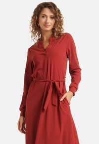Red long-sleeve dress with a v-neckline, belted waist, and soft, flowy fabric. Features deep side pockets and a relaxed fit.