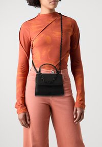Schwarze Lederhandtasche mit einer Kettenriemen, rechteckiger Form und Klappe vorne; getragen über einem orangen Langarmtop und pinken Hosen.