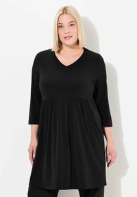 Mustat, polvipituiset tinukat, joissa on V-pääntie ja kolme neljänneshihaa. Valmistettu sileästä, joustavasta kankaasta; vyötäröllä on rypytyksiä.